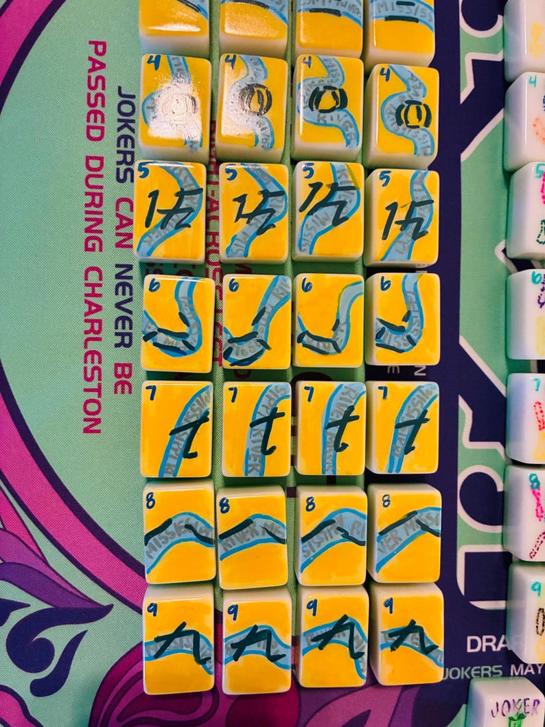 May include: A set of rectangular mahjong tiles with a yellow background and a blue river graphic. Each tile features a black number from 4 to 9 and the text Mississippi River. The tiles are arranged in rows on a mat with the text JOKERS CAN NEVER BE PASSED DURING CHARLESTON.