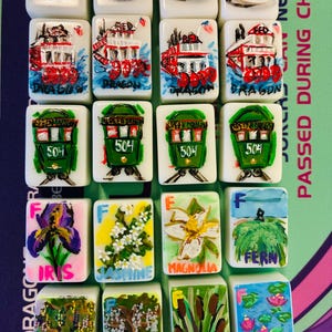 May include: A collection of twenty rectangular white tiles arranged in five rows of four. The tiles feature hand-painted illustrations including the Superdome, red and green streetcars, and various botanical subjects labeled with names like Iris, Jasmine, Magnolia, Fern, Oak, Cypress, Cattail, and Waterlily.