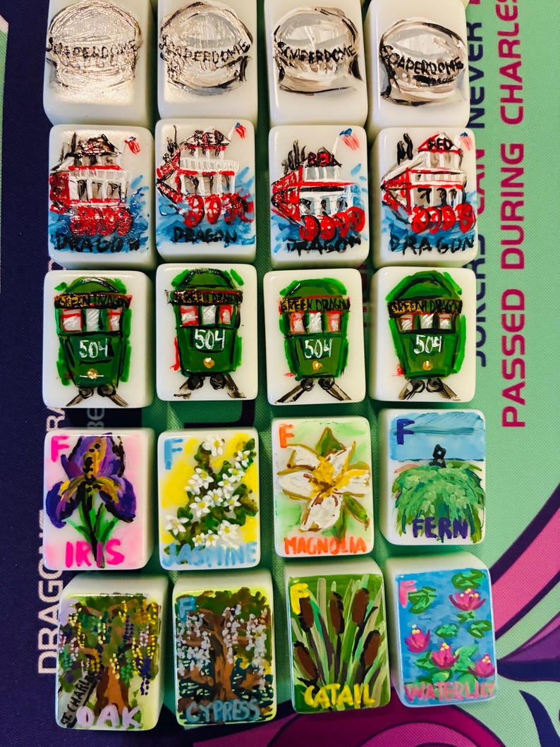 May include: A collection of twenty rectangular white tiles arranged in five rows of four. The tiles feature hand-painted illustrations including the Superdome, red and green streetcars, and various botanical subjects labeled with names like Iris, Jasmine, Magnolia, Fern, Oak, Cypress, Cattail, and Waterlily.