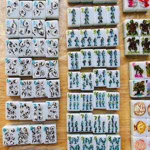 Puede incluir: Un conjunto de fichas de Mahjong blancas con diversos dise&ntilde;os pintados a mano. Las fichas presentan n&uacute;meros, criaturas marinas y escenas de la naturaleza en azul, rojo y amarillo. Est&aacute;n dispuestas sobre una superficie de madera marr&oacute;n claro.