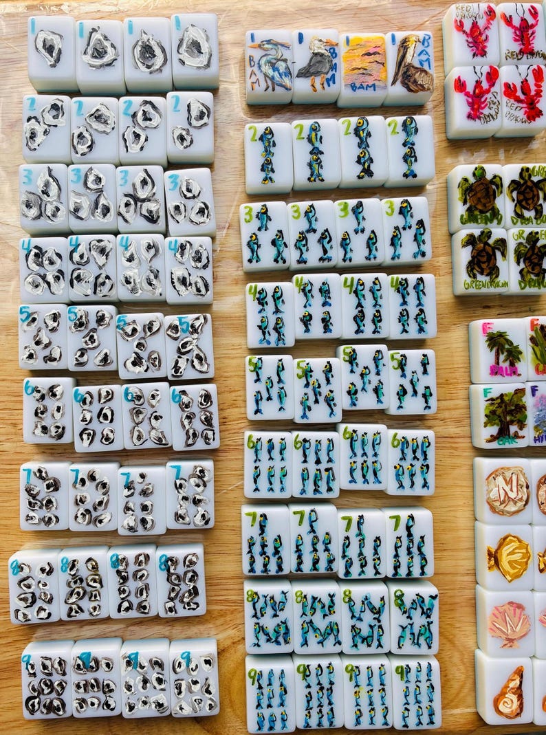 Puede incluir: Un conjunto de fichas de Mahjong blancas con diversos dise&ntilde;os pintados a mano. Las fichas presentan n&uacute;meros, criaturas marinas y escenas de la naturaleza en azul, rojo y amarillo. Est&aacute;n dispuestas sobre una superficie de madera marr&oacute;n claro.