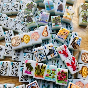 Puede incluir: Una colecci&oacute;n de fichas de Mahjong blancas, cada una pintada a mano con dise&ntilde;os &uacute;nicos. Las ilustraciones incluyen im&aacute;genes de tortugas, paisajes, flores y vida marina. Las fichas est&aacute;n dispersas, mostrando la variedad de ilustraciones.