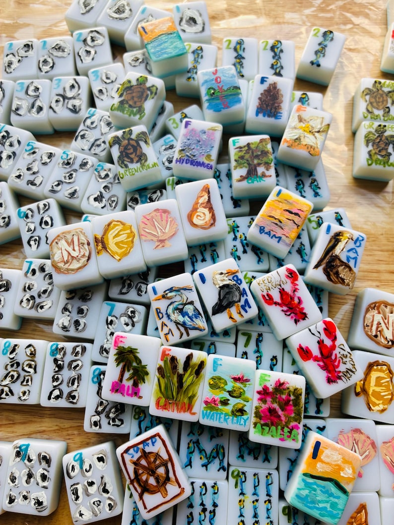 Puede incluir: Una colecci&oacute;n de fichas de Mahjong blancas, cada una pintada a mano con dise&ntilde;os &uacute;nicos. Las ilustraciones incluyen im&aacute;genes de tortugas, paisajes, flores y vida marina. Las fichas est&aacute;n dispersas, mostrando la variedad de ilustraciones.