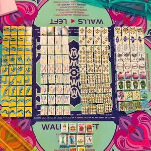 May include: A Mah Jongg game set featuring numerous white tiles with colorful illustrations of numbers, symbols, dragons, and flowers, arranged on a patterned green and purple mat with text instructions, accompanied by a translucent green ruler.