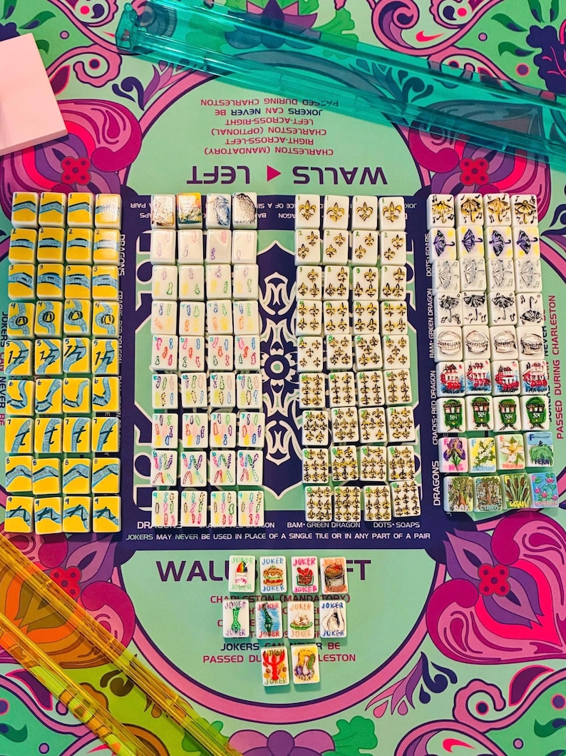 May include: A Mah Jongg game set featuring numerous white tiles with colorful illustrations of numbers, symbols, dragons, and flowers, arranged on a patterned green and purple mat with text instructions, accompanied by a translucent green ruler.