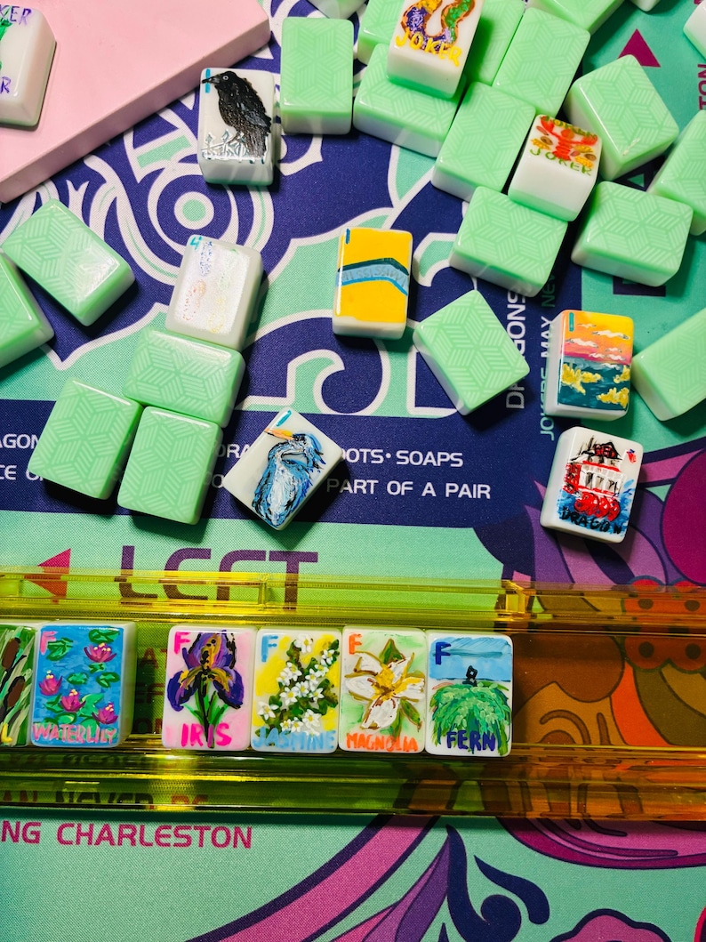 May include: A collection of rectangular mahjong tiles with light green backs featuring geometric patterns. Several tiles display hand-painted illustrations of flowers, birds, and landscapes, with labels including Waterlily, Iris, Jasmine, Magnolia, and Fern.