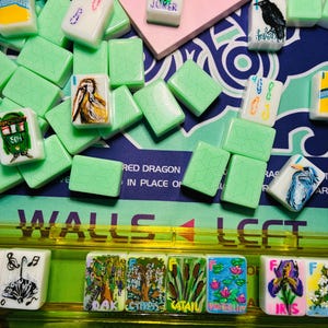 May include: A collection of light green mahjong tiles featuring various hand-painted illustrations including a joker, a crow, a pelican, a heron, and botanical scenes labeled oak, cypress, cattail, waterlily, and iris, arranged on a game mat.