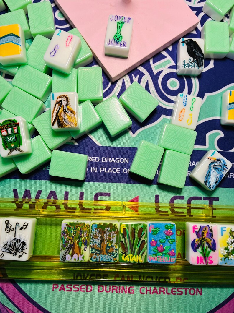 May include: A collection of light green mahjong tiles featuring various hand-painted illustrations including a joker, a crow, a pelican, a heron, and botanical scenes labeled oak, cypress, cattail, waterlily, and iris, arranged on a game mat.