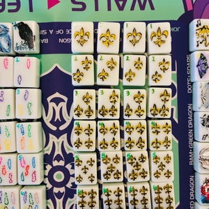 May include: A collection of white rectangular Mahjong tiles arranged in rows on a game mat. The tiles feature hand-painted yellow and black fleur-de-lis symbols in varying quantities, with small green numbers indicating the count of symbols per tile.
