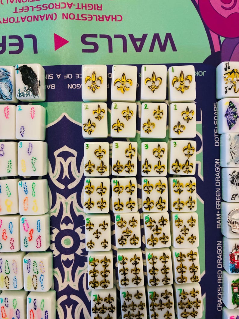 May include: A collection of white rectangular Mahjong tiles arranged in rows on a game mat. The tiles feature hand-painted yellow and black fleur-de-lis symbols in varying quantities, with small green numbers indicating the count of symbols per tile.