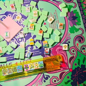May include: A Mahjong game set featuring light green tiles with various colorful illustrations and text, including Joker tiles. The tiles are arranged on a patterned game mat with purple, pink, and green floral designs and printed game instructions.