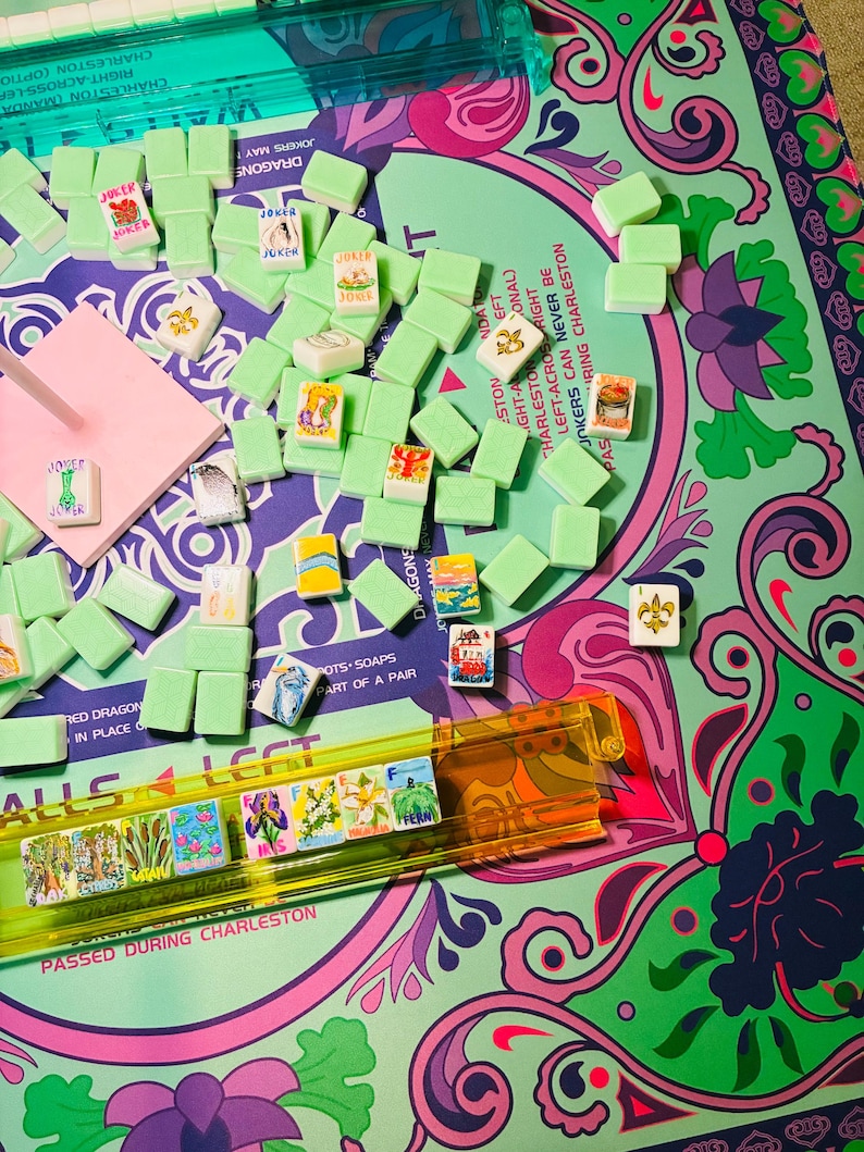 May include: A Mahjong game set featuring light green tiles with various colorful illustrations and text, including Joker tiles. The tiles are arranged on a patterned game mat with purple, pink, and green floral designs and printed game instructions.