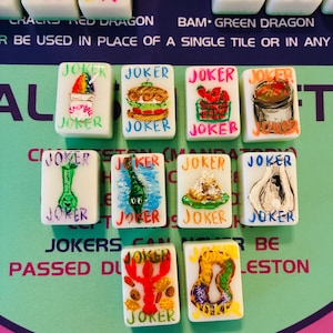 May include: A collection of ten white rectangular Mahjong tiles, each featuring the word JOKER in colorful lettering and a unique hand-painted illustration, including a snow cone, sandwich, strawberries, gumbo, alligator, crawfish, and king cake.