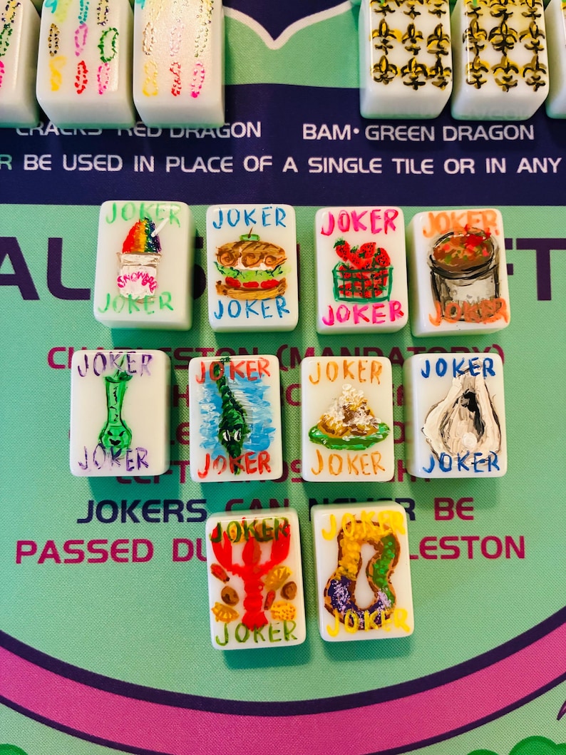 May include: A collection of ten white rectangular Mahjong tiles, each featuring the word JOKER in colorful lettering and a unique hand-painted illustration, including a snow cone, sandwich, strawberries, gumbo, alligator, crawfish, and king cake.