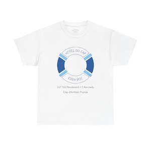May include: A white short-sleeve t-shirt featuring a blue lifebuoy graphic with the text HOTEL DU CAP EDEN-ROC. Below the graphic, the text reads 167-165 Boulevard J. F. Kennedy, Cap d'Antibes, France. The design uses shades of dark and light blue.
