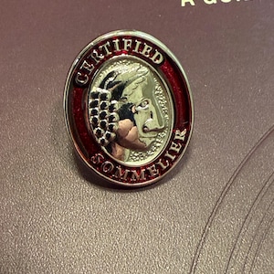 Puede incluir: Un pin de metal ovalado con un relieve plateado de una cabeza de perfil adornada con uvas. El borde es de esmalte rojo intenso con el texto CERTIFIED SOMMELIER en letras plateadas en relieve rodeando la figura central.