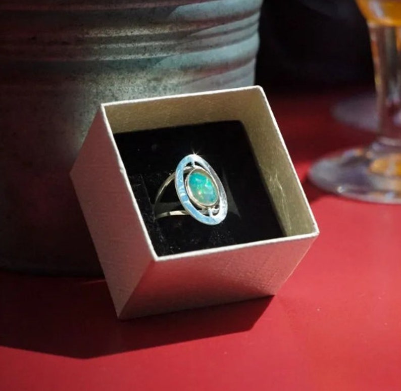 May include: A silver ring with a green opal stone set in a circular design. The ring is in a white box with a black interior.