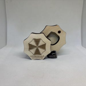 Puede incluir: Dos posavasos de madera con el logotipo de Umbrella Corporation. Un posavasos est&aacute; completo, el otro tiene un recorte circular. El logotipo es un emblema marr&oacute;n y blanco con el texto "UMBRELLA CORPORATION" debajo.