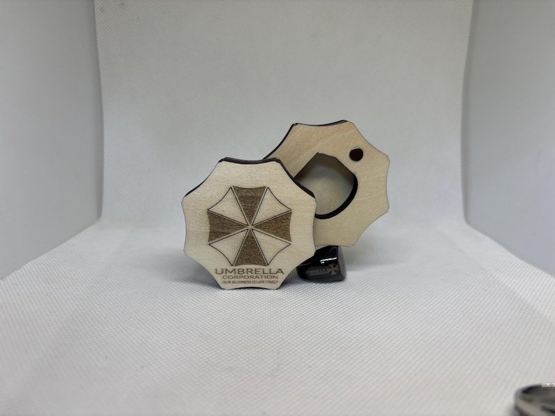 Puede incluir: Dos posavasos de madera con el logotipo de Umbrella Corporation. Un posavasos est&aacute; completo, el otro tiene un recorte circular. El logotipo es un emblema marr&oacute;n y blanco con el texto "UMBRELLA CORPORATION" debajo.