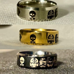 Könnte beinhalten: Drei verschiedene Metallringe mit gravierten Designs. Der erste Ring ist silber mit einem Totenkopf- und Stormtrooper-Design. Der zweite Ring ist gold mit einem Totenkopf- und Stormtrooper-Design. Der dritte Ring ist schwarz mit einem Totenkopf- und Stormtrooper-Design.