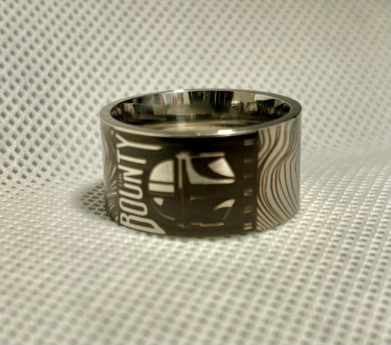 Pu&ograve; includere: Un anello d'argento con un design bianco e nero che presenta le parole "The Bounty" e un'immagine stilizzata di un'astronave. L'anello ha un motivo testurizzato e ondulato sul lato.