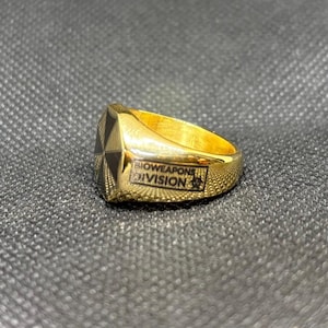 Puede incluir: Un anillo dorado con un dise&ntilde;o geom&eacute;trico negro y dorado en la cara. El anillo tiene la inscripci&oacute;n "BIOWEAPONS DIVISION". El anillo est&aacute; sobre un fondo texturizado gris oscuro.