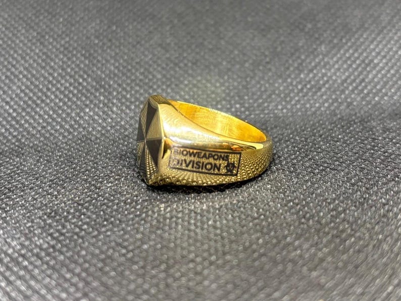 Puede incluir: Un anillo dorado con un dise&ntilde;o geom&eacute;trico negro y dorado en la cara. El anillo tiene la inscripci&oacute;n "BIOWEAPONS DIVISION". El anillo est&aacute; sobre un fondo texturizado gris oscuro.