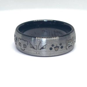 May include: A silver-colored ring with a black interior. The ring's exterior features engraved cartoon character faces. The ring is set against a white background, highlighting the details of the design.