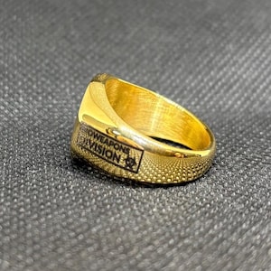 Puede incluir: Un anillo dorado con el texto "SHOWEAPONS DIVISION" en negro. El anillo tiene una superficie texturizada y un emblema rectangular con un s&iacute;mbolo de peligro biol&oacute;gico. El anillo est&aacute; sobre un fondo gris oscuro.