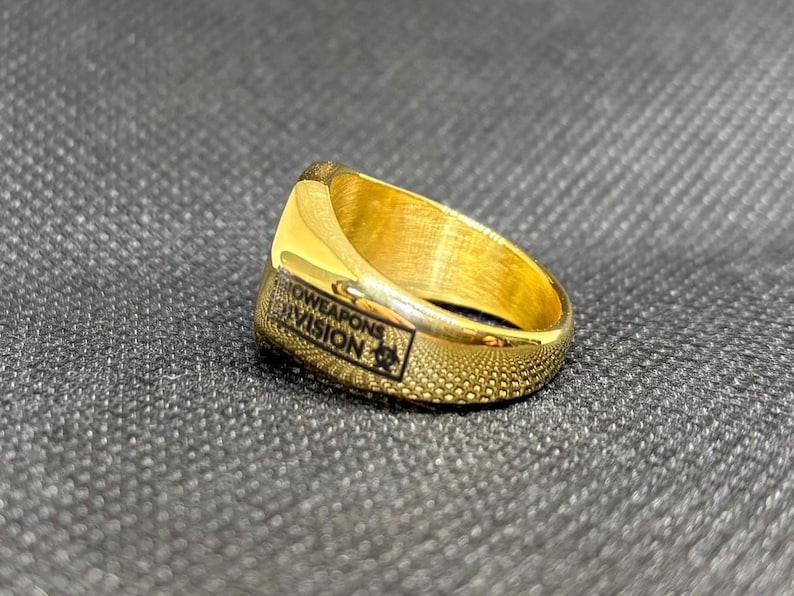 Puede incluir: Un anillo dorado con el texto "SHOWEAPONS DIVISION" en negro. El anillo tiene una superficie texturizada y un emblema rectangular con un s&iacute;mbolo de peligro biol&oacute;gico. El anillo est&aacute; sobre un fondo gris oscuro.