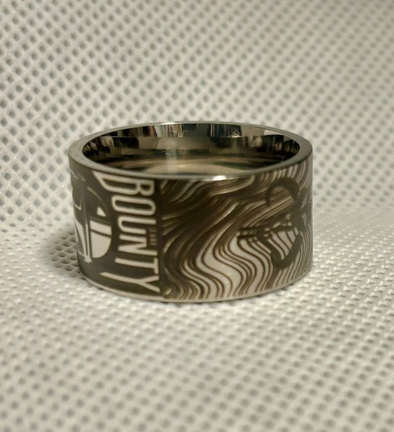 Pu&ograve; includere: Un anello d'argento con un design bianco e nero che presenta le parole "BOUNTY HUNTER" e un'immagine stilizzata di un'astronave.