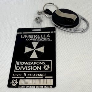 Puede incluir: Tarjeta de identificaci&oacute;n negra de Umbrella Corporation con un llavero plateado y un carrete retr&aacute;ctil. La tarjeta presenta el logotipo de Umbrella Corporation, el texto "BIOWEAPONS DIVISION LEVEL 5 CLEARANCE" y un c&oacute;digo de acceso.