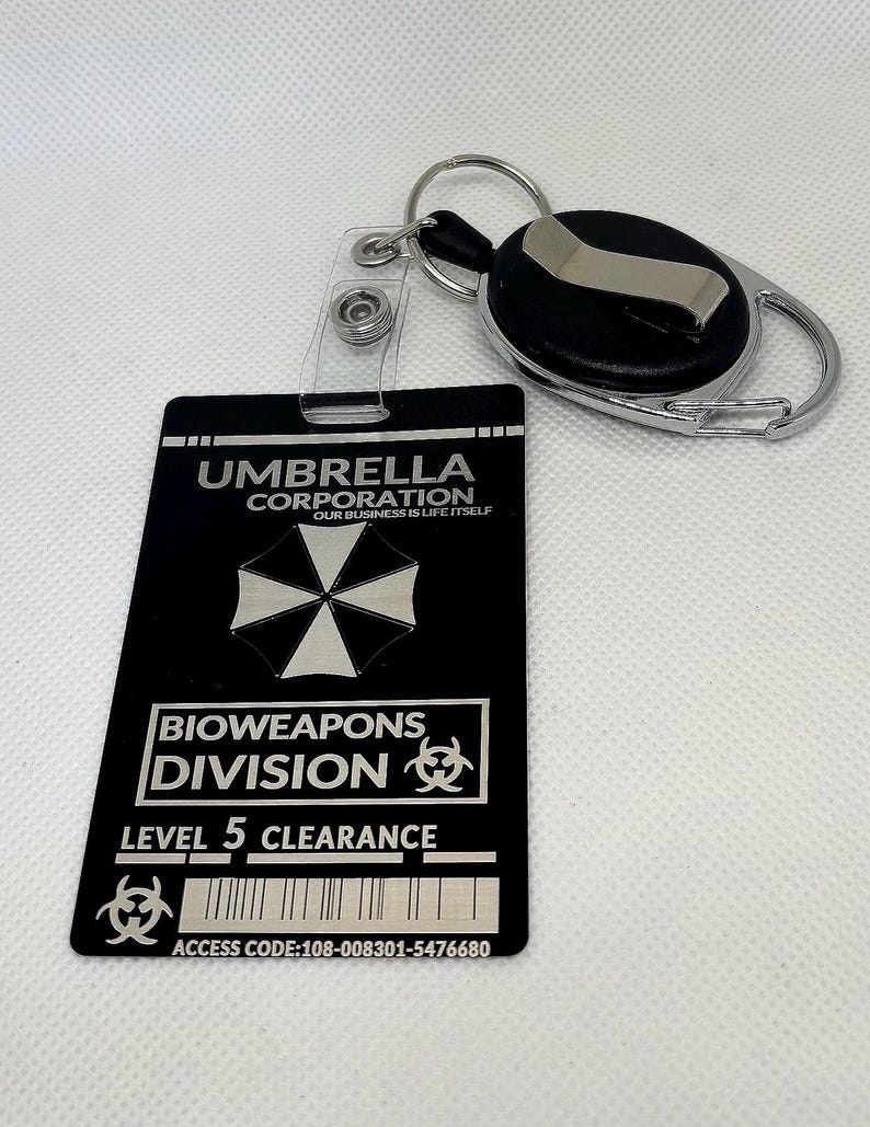 Puede incluir: Tarjeta de identificaci&oacute;n negra de Umbrella Corporation con un llavero plateado y un carrete retr&aacute;ctil. La tarjeta presenta el logotipo de Umbrella Corporation, el texto "BIOWEAPONS DIVISION LEVEL 5 CLEARANCE" y un c&oacute;digo de acceso.