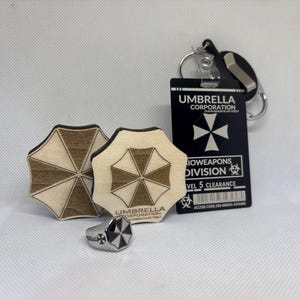 Puede incluir: Un surtido de productos de Umbrella Corporation, que incluye dos posavasos de madera, un anillo plateado y una tarjeta de identificaci&oacute;n negra con un llavero. Los posavasos y el anillo presentan el logotipo de Umbrella Corporation. La tarjeta de identificaci&oacute;n dice "Umbrella Corporation" y "Bioweapons Division."
