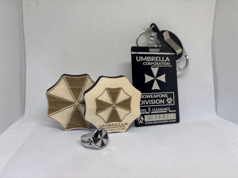 Puede incluir: Un surtido de productos de Umbrella Corporation, que incluye dos posavasos de madera, un anillo plateado y una tarjeta de identificaci&oacute;n negra con un llavero. Los posavasos y el anillo presentan el logotipo de Umbrella Corporation. La tarjeta de identificaci&oacute;n dice "Umbrella Corporation" y "Bioweapons Division."