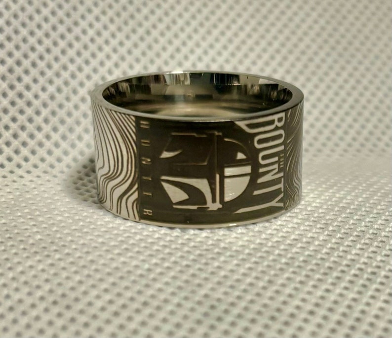 Pu&ograve; includere: Un anello d'argento con un designo in bianco e nero di un'astronave con la parola "BOUNTY" incisa sul lato.