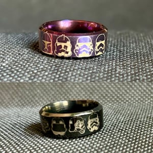 Könnte beinhalten: Zwei schwarze und goldene Ringe mit einem Design aus fünf Star Wars Stormtrooper-Helmen. Der obere Ring ist lila.
