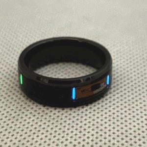 Op de afbeelding: Een zwarte ring met groene en blauwe glow-in-the-dark accenten. De ring is gemaakt van keramiek en heeft een gladde, gepolijste afwerking.