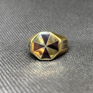 Puede incluir: Un anillo dorado con un dise&ntilde;o geom&eacute;trico. El anillo presenta un oct&aacute;gono central con segmentos alternos de color p&uacute;rpura oscuro y marr&oacute;n rojizo. La banda tiene texto grabado. El anillo est&aacute; sobre una superficie gris texturizada.