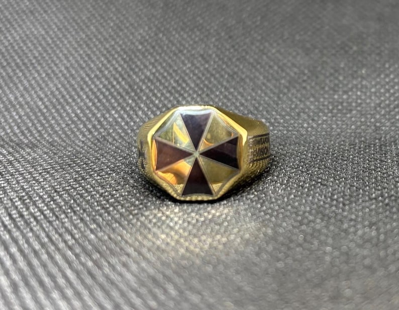 Puede incluir: Un anillo dorado con un dise&ntilde;o geom&eacute;trico. El anillo presenta un oct&aacute;gono central con segmentos alternos de color p&uacute;rpura oscuro y marr&oacute;n rojizo. La banda tiene texto grabado. El anillo est&aacute; sobre una superficie gris texturizada.