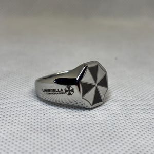 Puede incluir: Anillo plateado con un logotipo de paraguas en blanco y negro en la parte superior. El anillo tiene la inscripci&oacute;n "UMBRELLA CORPORATION" y un logotipo a juego en el lateral. Sobre un fondo blanco texturizado.