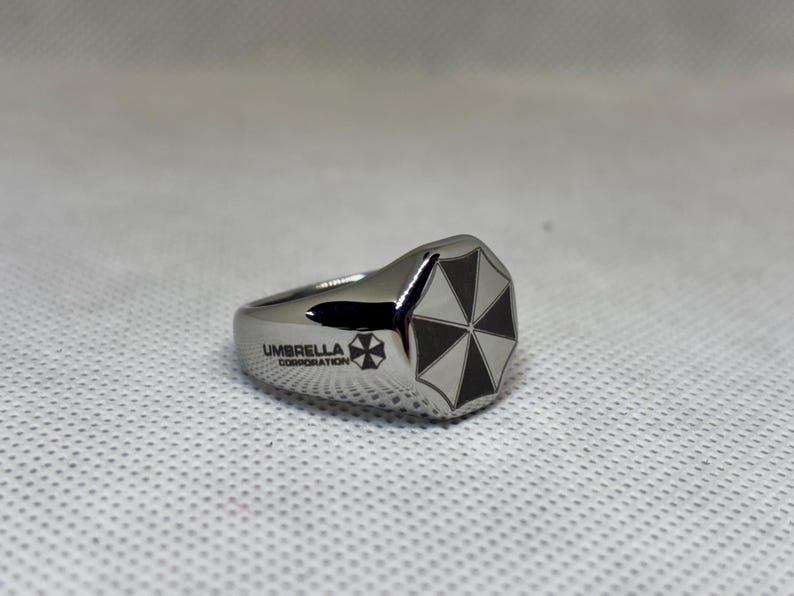 Puede incluir: Anillo plateado con un logotipo de paraguas en blanco y negro en la parte superior. El anillo tiene la inscripci&oacute;n "UMBRELLA CORPORATION" y un logotipo a juego en el lateral. Sobre un fondo blanco texturizado.
