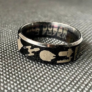 May include: A black stainless steel ring with a silver engraved design of Star Wars characters, including the Millennium Falcon, X-Wing, and a TIE fighter. The ring also has the text "Star Wars" engraved on the inside.