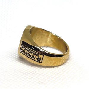 Puede incluir: Anillo dorado con una cara rectangular. La cara del anillo tiene las palabras "BIOWEAPONS DIVISION" y un s&iacute;mbolo de peligro biol&oacute;gico en negro. El anillo est&aacute; sobre un fondo blanco.