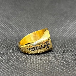 Puede incluir: Un anillo dorado con el texto "UMBRELLA CORPORATION" y un logotipo en blanco y negro. El anillo tiene una banda texturizada y una parte superior plana con el logotipo. El anillo est&aacute; sobre un fondo gris oscuro.