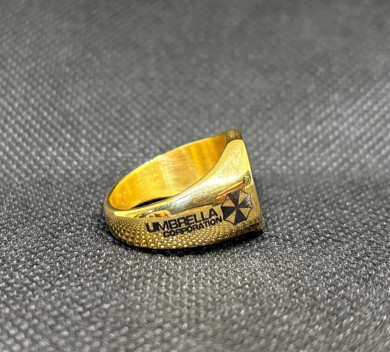 Puede incluir: Un anillo dorado con el texto "UMBRELLA CORPORATION" y un logotipo en blanco y negro. El anillo tiene una banda texturizada y una parte superior plana con el logotipo. El anillo est&aacute; sobre un fondo gris oscuro.