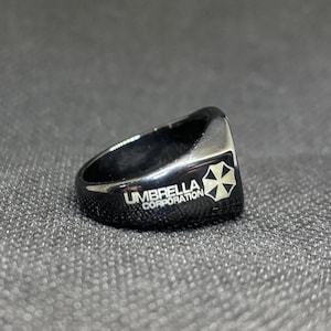 Puede incluir: Anillo negro con el texto "UMBRELLA CORPORATION" y un logotipo blanco y negro. El anillo es de estilo sello, con una parte superior plana y bordes redondeados. El anillo est&aacute; hecho de un material brillante y reflectante.
