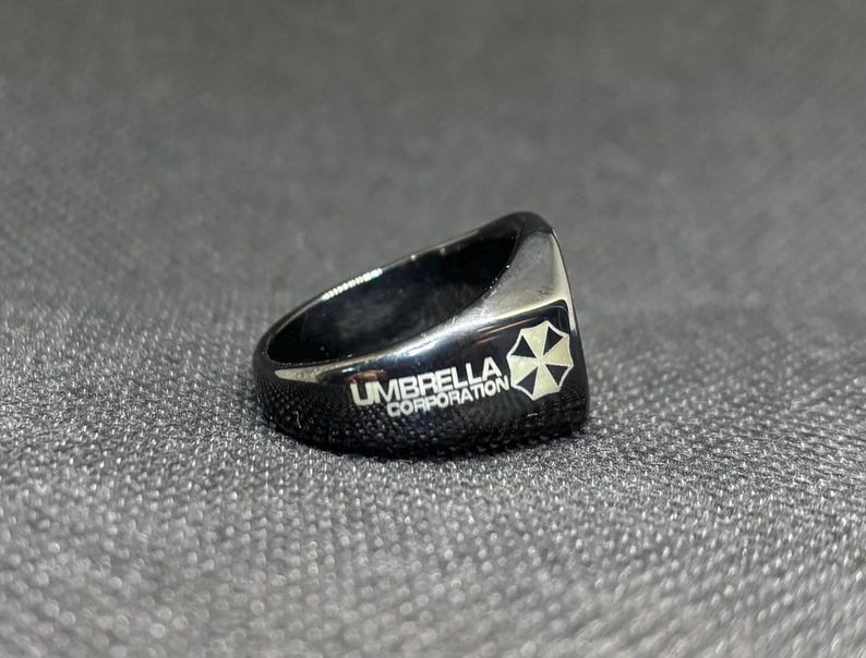 Puede incluir: Anillo negro con el texto "UMBRELLA CORPORATION" y un logotipo blanco y negro. El anillo es de estilo sello, con una parte superior plana y bordes redondeados. El anillo est&aacute; hecho de un material brillante y reflectante.