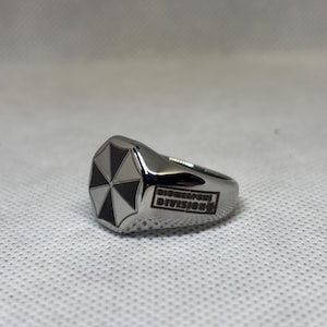 Puede incluir: Un anillo plateado con un logotipo de paraguas en blanco y negro en la cara. El anillo tiene las palabras "Biohazard Division" grabadas en el lateral. El anillo est&aacute; sobre un fondo blanco texturizado.