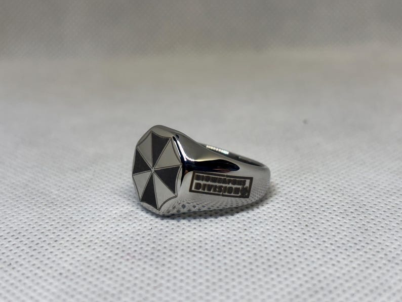 Puede incluir: Un anillo plateado con un logotipo de paraguas en blanco y negro en la cara. El anillo tiene las palabras "Biohazard Division" grabadas en el lateral. El anillo est&aacute; sobre un fondo blanco texturizado.